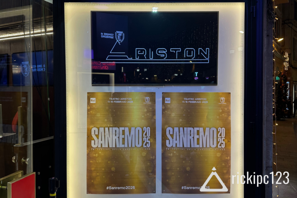 Vetrina del Teatro Ariston di Sanremo con il logo illuminato in alto e due poster ufficiali del Festival di Sanremo 2025, con dettagli sull’evento dal 11 al 15 febbraio. L’immagine include il watermark “rickipc123”.