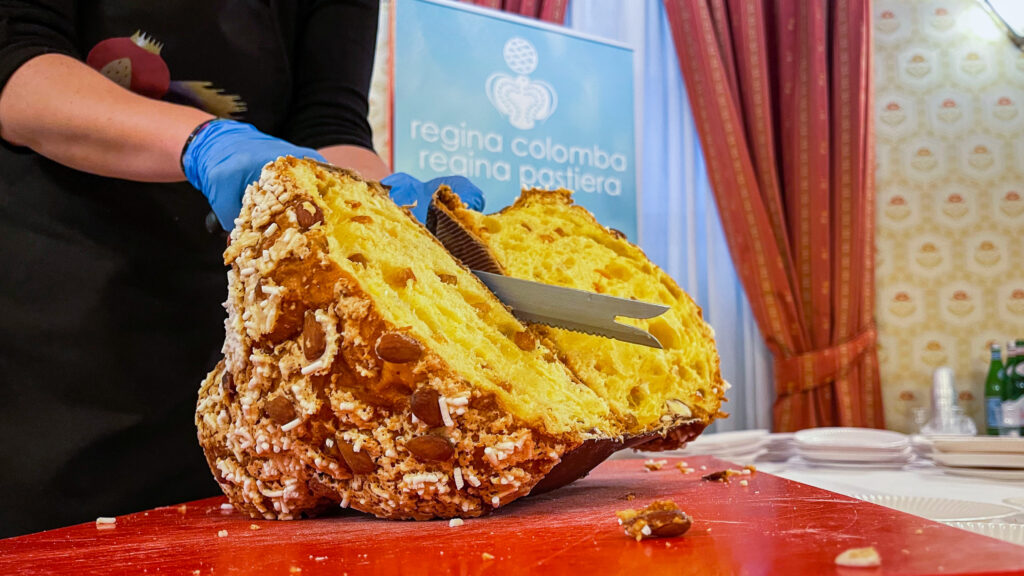 Colomba artigianale tagliata durante l’evento Regina Colomba Regina Pastiera, con il logo della manifestazione sullo sfondo.